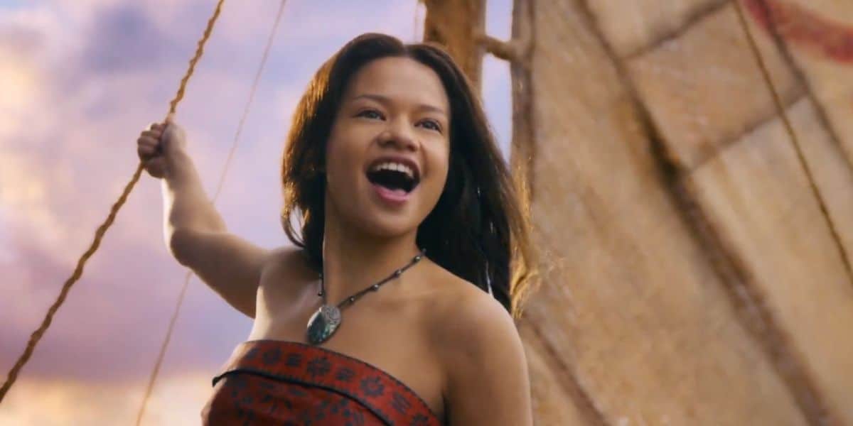 A young woman with long dark hair, wearing a patterned strapless dress and a necklace, stands on a boat holding a rope and smiling joyfully with the sail and sky in the background, evoking Moana live-action adventure vibes.