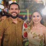 Homem e mulher sorriem lado a lado em evento noturno com luzes coloridas ao fundo. Ele usa camisa xadrez amarela e preta; ela veste traje típico de festa junina com bordados florais em vermelho, dourado e azul.
