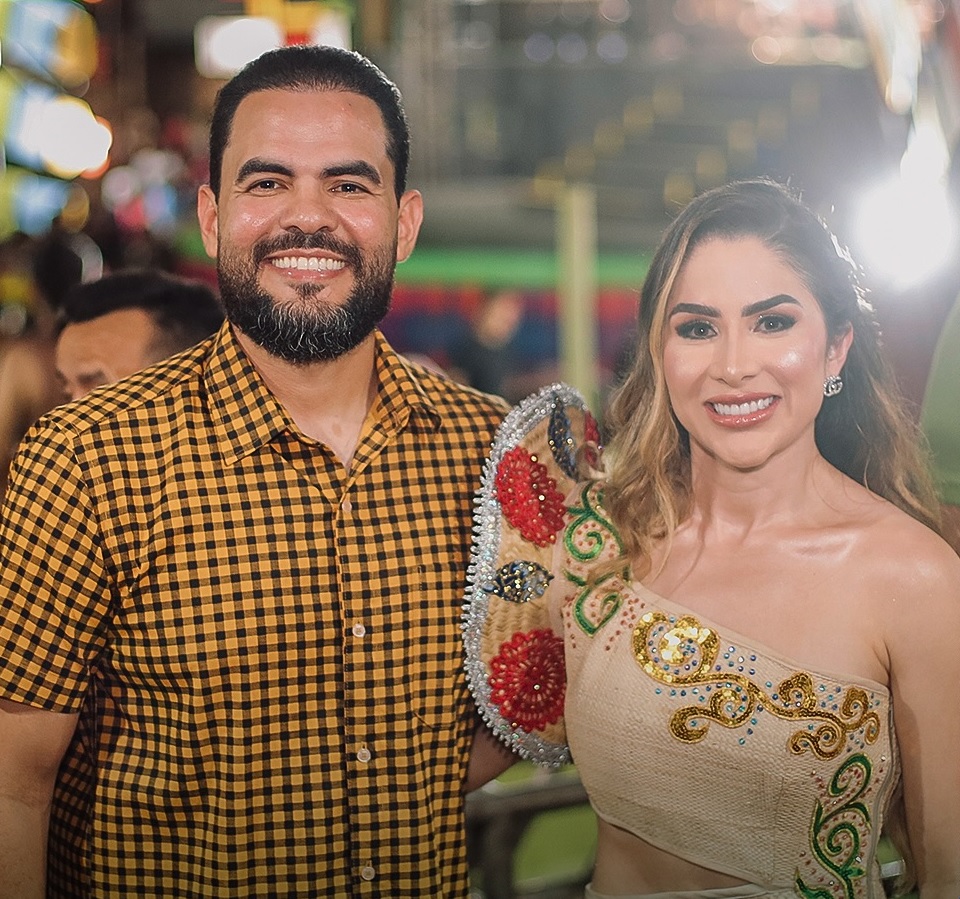 Homem e mulher sorriem lado a lado em evento noturno com luzes coloridas ao fundo. Ele usa camisa xadrez amarela e preta; ela veste traje típico de festa junina com bordados florais em vermelho, dourado e azul.
