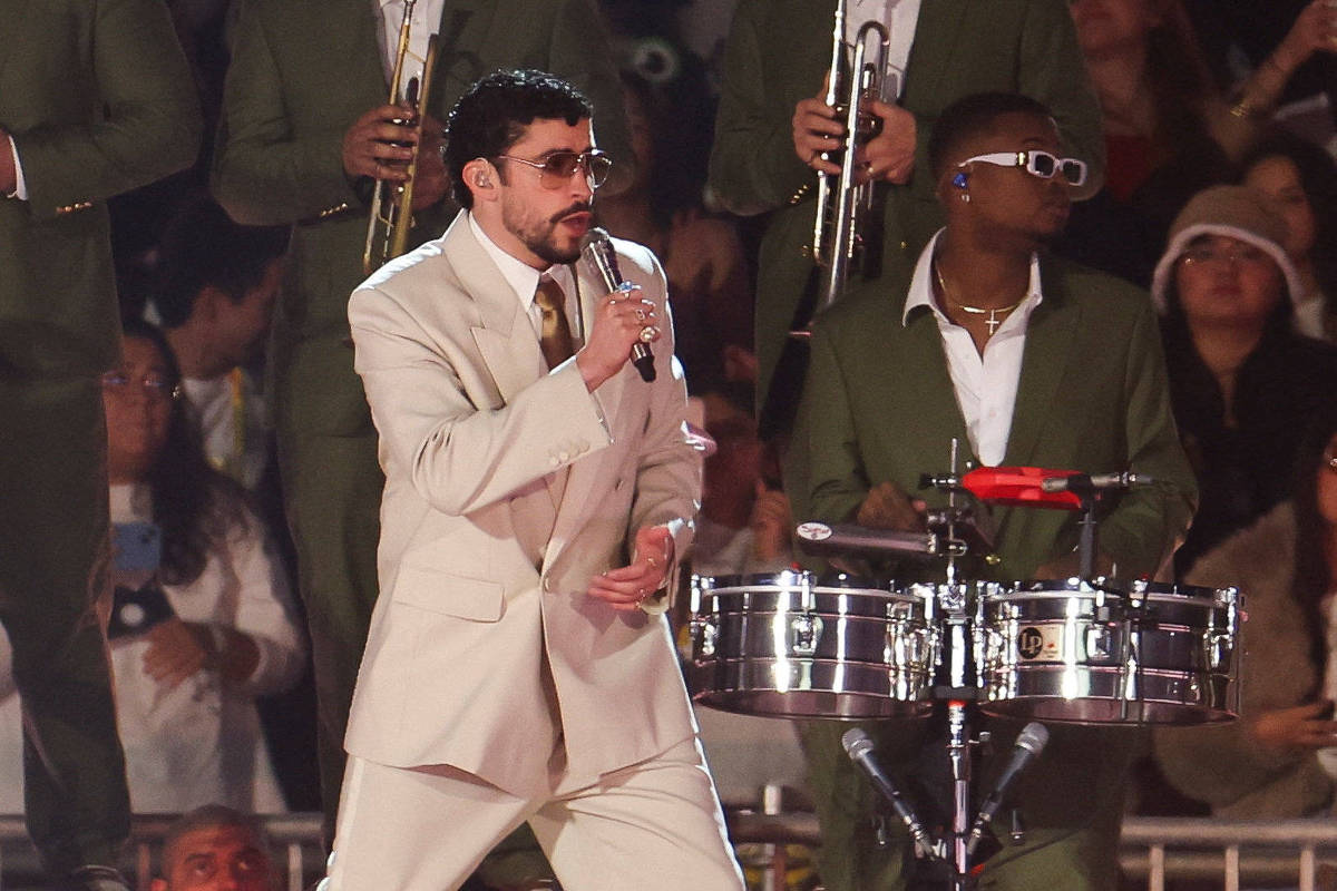 Cantor de terno bege claro e óculos escuros segura microfone e canta. Ao lado, percussionista com óculos brancos toca timbales, ambos em palco com músicos ao fundo e público visível.